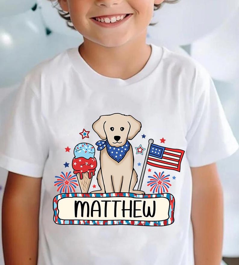 Pu&ograve; includere: Maglietta bianca con un design patriottico rosso, bianco e blu con un cane, un cono gelato e fuochi d'artificio. Il testo "MATTHEW" &egrave; stampato su uno stendardo.