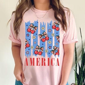 May include: Pink t-shirt with a blue and red graphic of cherries and stars with the word "AMERICA" printed below.
