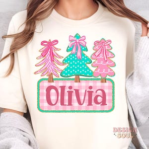 May include: Cream-colored t-shirt featuring three Christmas trees with pink bows and gold accents. The center tree is teal with white polka dots. Below, a pink and white plaid rectangle displays the name "Olivia".