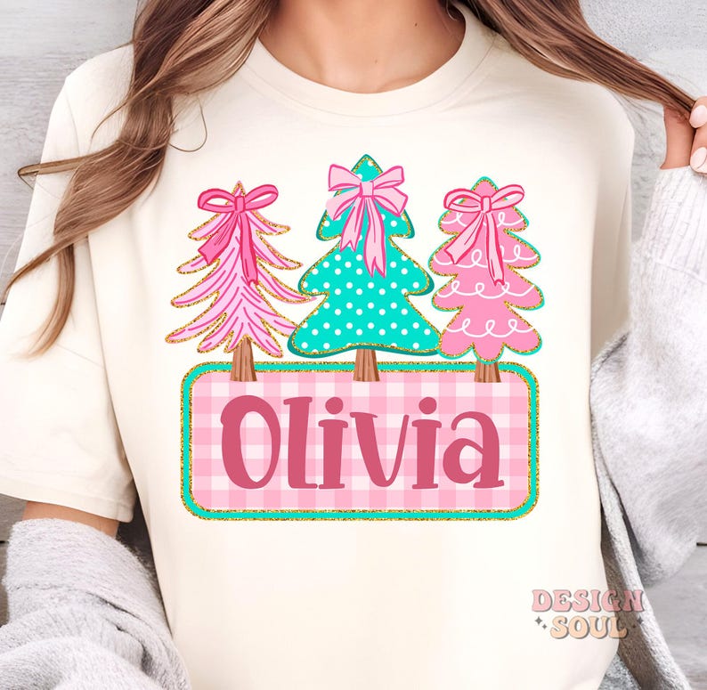 May include: Cream-colored t-shirt featuring three Christmas trees with pink bows and gold accents. The center tree is teal with white polka dots. Below, a pink and white plaid rectangle displays the name "Olivia".