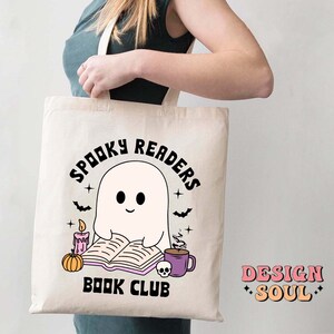 May include: A white tote bag with a black and white illustration of a ghost reading a book. The text on the bag reads "Spooky Readers Book Club".