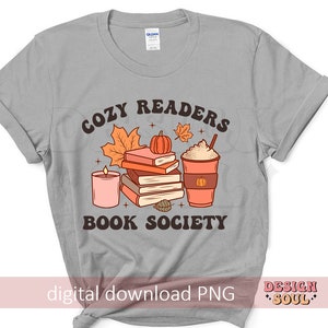 May include: Gray t-shirt with a graphic design of a stack of books, a candle, a pumpkin, and a cup of coffee. The text "Cozy Readers Book Society" is printed on the shirt.