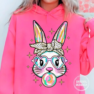 May include: Pink hoodie with a graphic of a cartoon bunny wearing heart-shaped sunglasses and a polka dot headband. The bunny is holding a colorful lollipop.