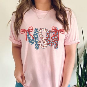 May include: A pink t-shirt with the word "USA" in red, white, and blue patterned letters. The letters are decorated with red bows.