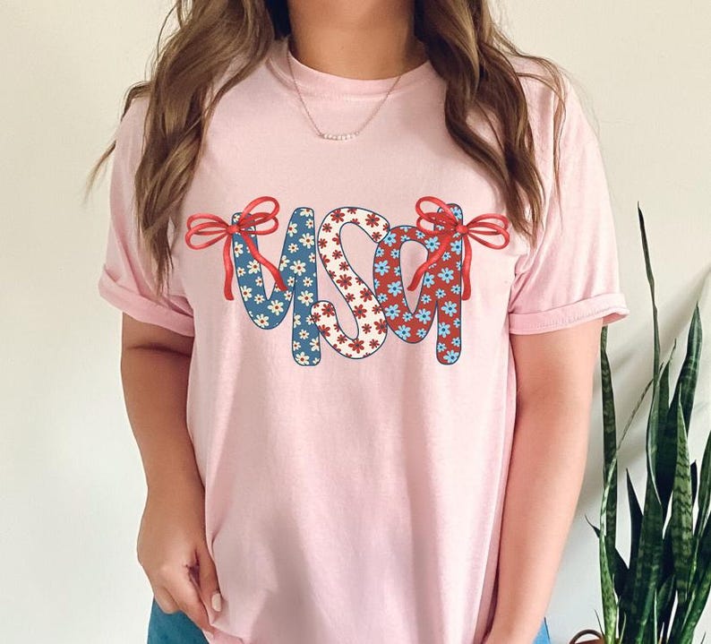 May include: A pink t-shirt with the word "USA" in red, white, and blue patterned letters. The letters are decorated with red bows.