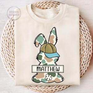 May include: A beige sweatshirt with a camouflage bunny rabbit wearing a green and blue baseball cap. The bunny has a name tag that says "MATTHEW".