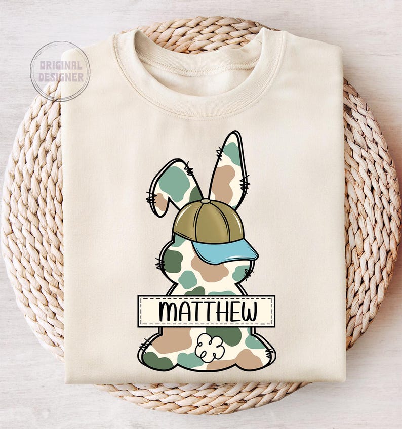 May include: A beige sweatshirt with a camouflage bunny rabbit wearing a green and blue baseball cap. The bunny has a name tag that says "MATTHEW".