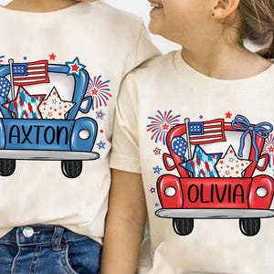 May include: Two cream-colored t-shirts with matching patriotic designs. One shirt features a blue truck with the name "AXTON" and the other a red truck with the name "OLIVIA". Both designs include an American flag, stars, and fireworks.