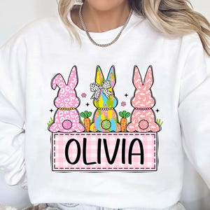 May include: White sweatshirt with a pink, yellow, and blue bunny design. The bunnies are holding carrots and have a pink bow. The text "Olivia" is written in a pink checkered box.