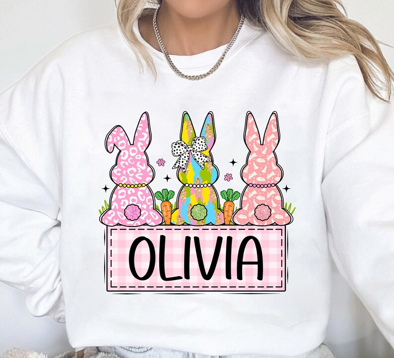 May include: White sweatshirt with a pink, yellow, and blue bunny design. The bunnies are holding carrots and have a pink bow. The text "Olivia" is written in a pink checkered box.