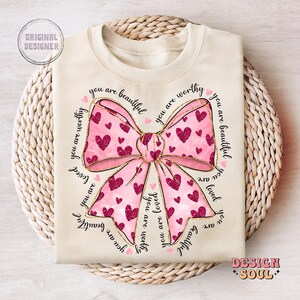 May include: A cream-colored t-shirt with a pink bow and the text "You are beautiful, you are worthy, you are loved" repeated around the bow.