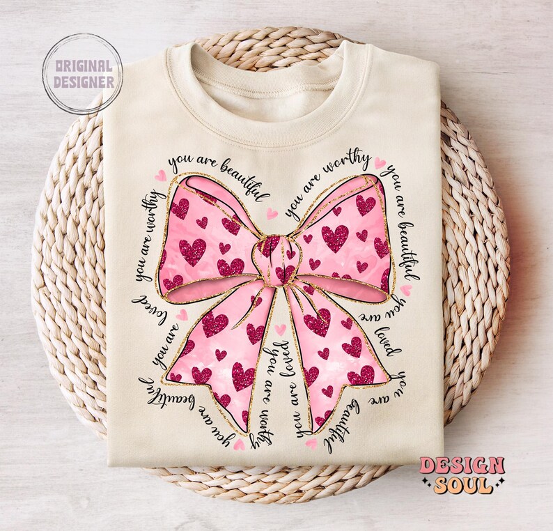 May include: A cream-colored t-shirt with a pink bow and the text "You are beautiful, you are worthy, you are loved" repeated around the bow.