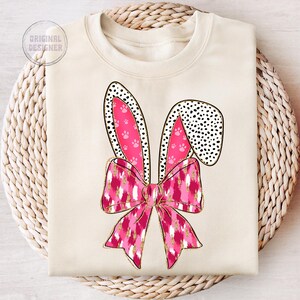 May include: A cream-coloured sweatshirt featuring a pink and white bunny ear design. The bunny ears have a pink and white polka dot pattern and pink paw prints. The bunny ears are tied together with a pink and white bow with a gold glitter pattern.
