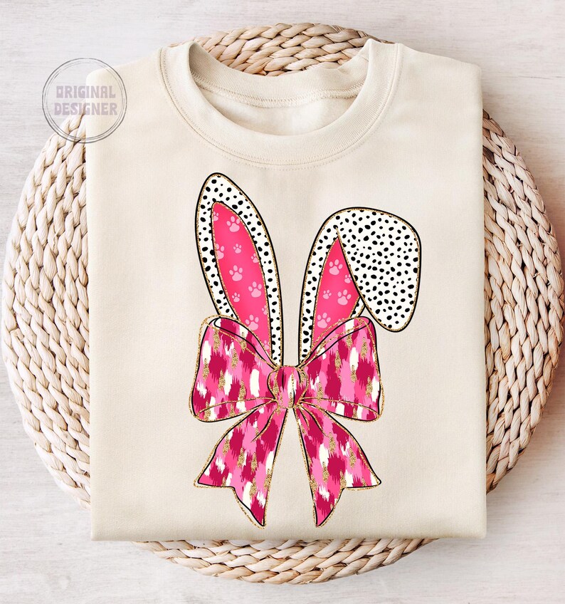 May include: A cream-coloured sweatshirt featuring a pink and white bunny ear design. The bunny ears have a pink and white polka dot pattern and pink paw prints. The bunny ears are tied together with a pink and white bow with a gold glitter pattern.