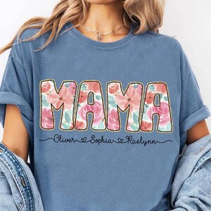 May include: A blue t-shirt with the word "MAMA" in gold letters with a floral pattern. The names "Oliver", "Sophia", and "Raelynn" are written below the word "MAMA" with hearts between each name.