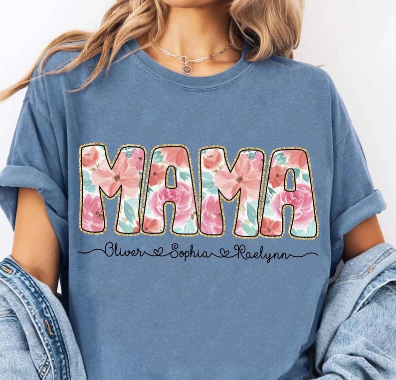 May include: A blue t-shirt with the word "MAMA" in gold letters with a floral pattern. The names "Oliver", "Sophia", and "Raelynn" are written below the word "MAMA" with hearts between each name.
