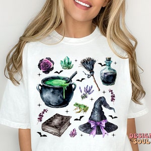 May include: White t-shirt with a watercolor design of a witch's cauldron, a broom, a potion bottle, a frog, a hat, crystals, and flowers. The design is in shades of green, black, purple, and white.