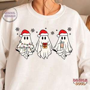 May include: White sweatshirt with a Christmas-themed design featuring three ghosts wearing Santa hats. The ghosts are holding a gift, a cup of coffee, and Christmas lights.