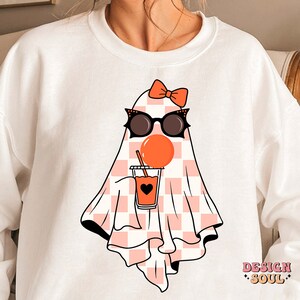 May include: White sweatshirt with a pink and white checkered ghost wearing sunglasses and a pink bow. The ghost is holding a pink cup with a black heart and orange liquid. The text "DESIGN SOUL" is printed on the bottom right corner of the sweatshirt.