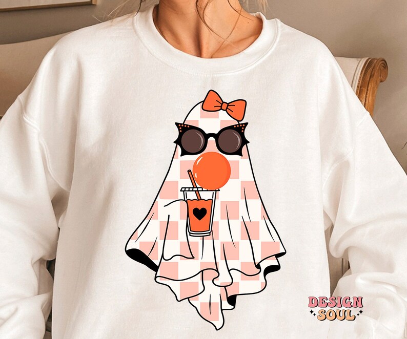 May include: White sweatshirt with a pink and white checkered ghost wearing sunglasses and a pink bow. The ghost is holding a pink cup with a black heart and orange liquid. The text "DESIGN SOUL" is printed on the bottom right corner of the sweatshirt.
