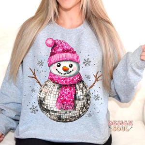 May include: A gray sweatshirt with a pink and silver disco ball snowman design. The snowman is wearing a pink hat and scarf. The text "Design Soul" is printed on the bottom right corner of the sweatshirt.