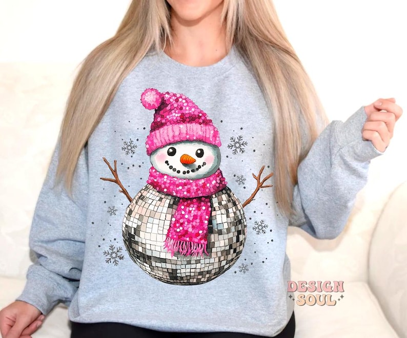 May include: A gray sweatshirt with a pink and silver disco ball snowman design. The snowman is wearing a pink hat and scarf. The text "Design Soul" is printed on the bottom right corner of the sweatshirt.