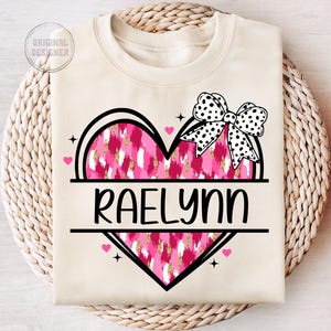May include: A cream-colored sweatshirt with a pink and gold heart design. The heart has a black outline and a black and white polka dot bow on top. The name "RAELYN" is written in black letters below the heart.
