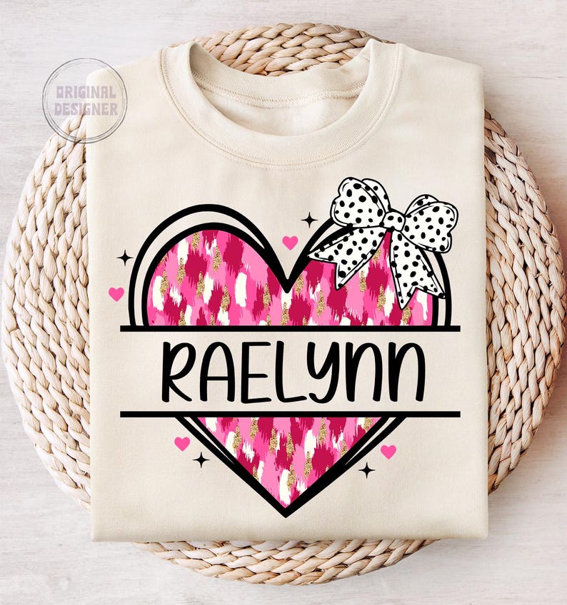May include: A cream-colored sweatshirt with a pink and gold heart design. The heart has a black outline and a black and white polka dot bow on top. The name "RAELYN" is written in black letters below the heart.
