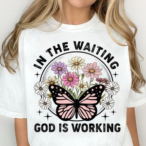 May include: White t-shirt with a graphic design featuring a butterfly with pink and black wings, surrounded by flowers. The text "In the waiting, God is working" is printed in black.