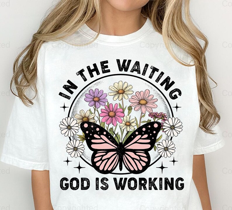 May include: White t-shirt with a graphic design featuring a butterfly with pink and black wings, surrounded by flowers. The text "In the waiting, God is working" is printed in black.