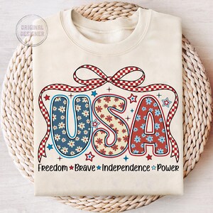 May include: A beige sweatshirt with a red, white, and blue USA graphic. The graphic features a red and white checkered bow, stars, and flowers. The text "Freedom Brave Independence Power" is printed below the graphic.