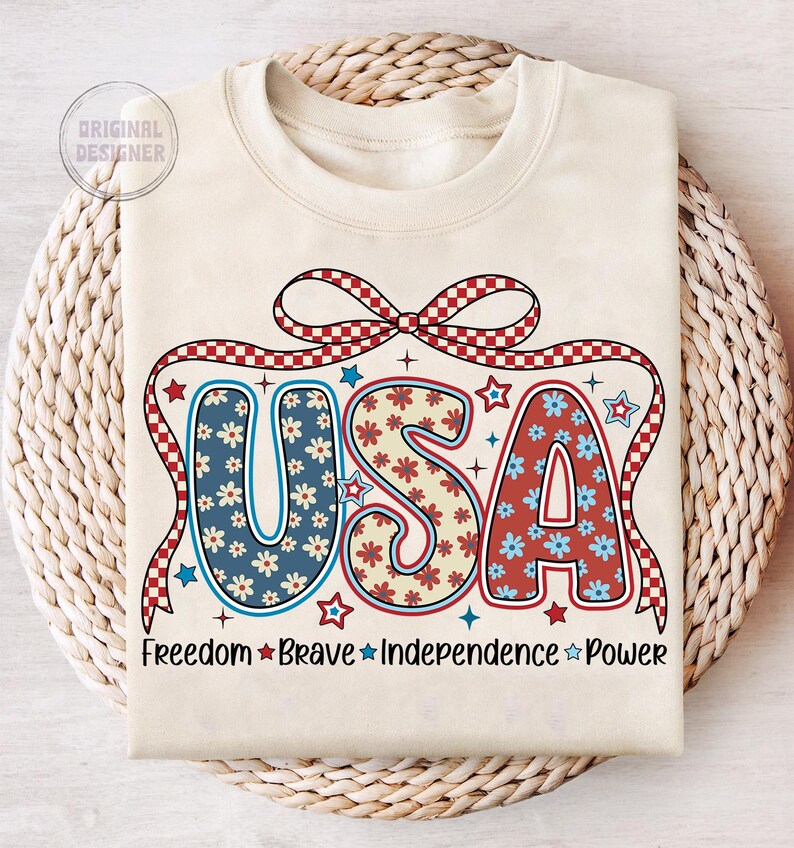 May include: A beige sweatshirt with a red, white, and blue USA graphic. The graphic features a red and white checkered bow, stars, and flowers. The text "Freedom Brave Independence Power" is printed below the graphic.