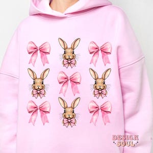 May include: Pink hoodie with a repeating pattern of brown bunnies wearing pink bows. The hoodie has a large hood and long sleeves.