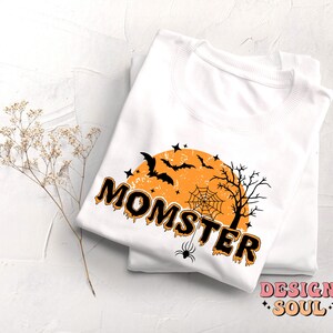 May include: White t-shirt with a black and orange graphic design that says "Momster" with a spider, bats, and a moon in the background.