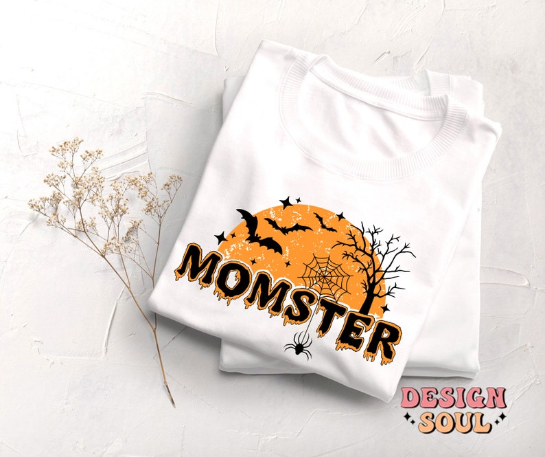 May include: White t-shirt with a black and orange graphic design that says "Momster" with a spider, bats, and a moon in the background.