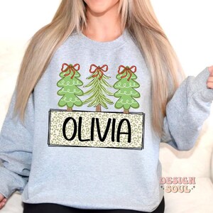 May include: A light gray sweatshirt with a design of three green Christmas trees with red bows and the name "Olivia" in a brown leopard print.