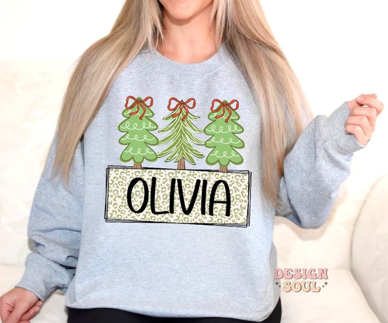 May include: A light gray sweatshirt with a design of three green Christmas trees with red bows and the name "Olivia" in a brown leopard print.