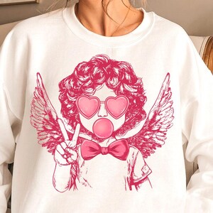May include: White sweatshirt featuring a pink graphic of a cherub with angel wings wearing heart-shaped sunglasses and blowing a bubble. The cherub is giving the peace sign.