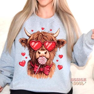 May include: A gray sweatshirt with a graphic of a Highland cow wearing heart-shaped sunglasses and a bow tie. The cow is holding a red and white lollipop. The text "DESIGN SOUL" is printed on the bottom right corner of the sweatshirt.