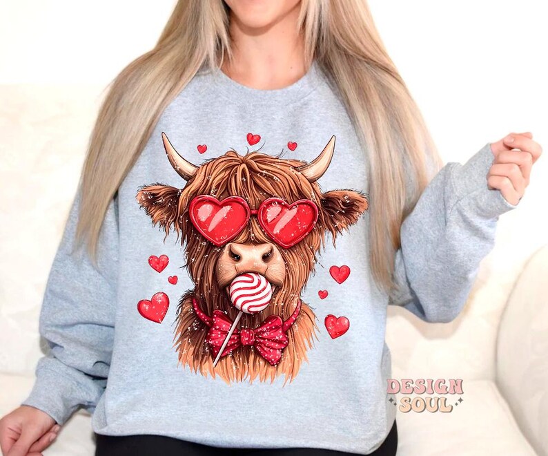 May include: A gray sweatshirt with a graphic of a Highland cow wearing heart-shaped sunglasses and a bow tie. The cow is holding a red and white lollipop. The text "DESIGN SOUL" is printed on the bottom right corner of the sweatshirt.
