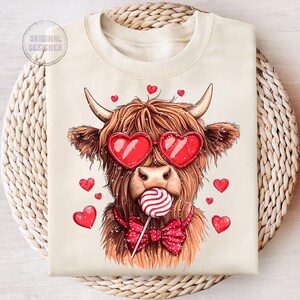 May include: A beige sweatshirt with a cartoon illustration of a Highland cow wearing heart-shaped sunglasses and a red bow tie. The cow is holding a red and white lollipop in its mouth. There are red hearts scattered around the cow.