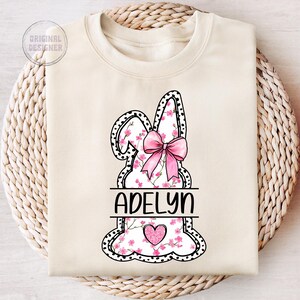 Pu&ograve; includere: Una felpa bianca con un design a forma di coniglio rosa. Il coniglio &egrave; contornato di nero con pois bianchi e ha un fiocco rosa. Il coniglio &egrave; riempito con un motivo floreale rosa. Il nome "Adelyn" &egrave; scritto in lettere nere sul corpo del coniglio. Un cuore rosa con glitter si trova al centro del coniglio.