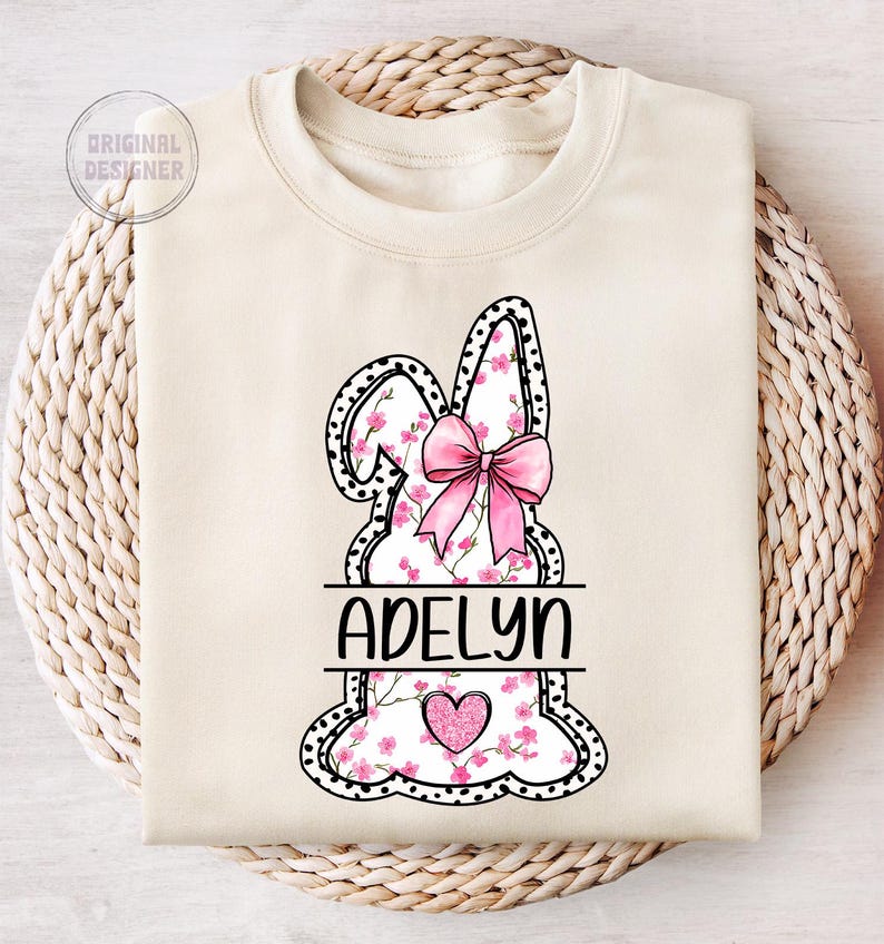 Pu&ograve; includere: Una felpa bianca con un design a forma di coniglio rosa. Il coniglio &egrave; contornato di nero con pois bianchi e ha un fiocco rosa. Il coniglio &egrave; riempito con un motivo floreale rosa. Il nome "Adelyn" &egrave; scritto in lettere nere sul corpo del coniglio. Un cuore rosa con glitter si trova al centro del coniglio.
