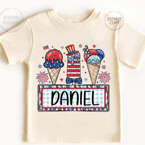 May include: Cream-colored t-shirt featuring a patriotic ice cream design with red, white, and blue colors. The design includes ice cream cones and popsicles, a small American flag, and the name "DANIEL" in a rectangular frame.