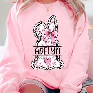 Pu&ograve; includere: Felpa rosa con un grafico di coniglio bianco e nero. Il coniglio ha fiori rosa e un fiocco rosa. Il nome "Adelyn" &egrave; scritto sotto il coniglio.