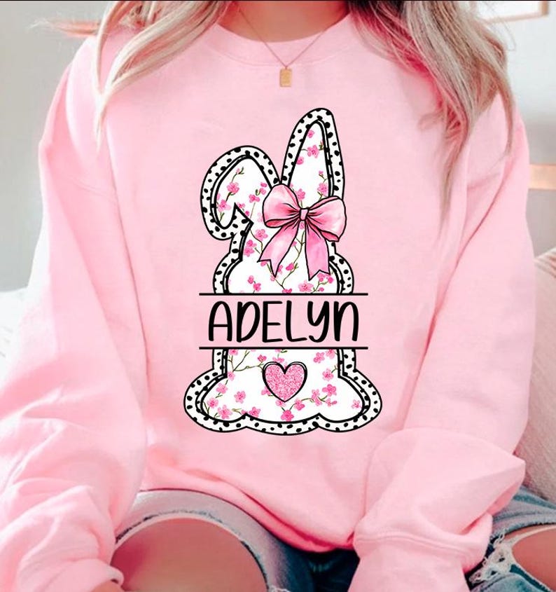 Pu&ograve; includere: Felpa rosa con un grafico di coniglio bianco e nero. Il coniglio ha fiori rosa e un fiocco rosa. Il nome "Adelyn" &egrave; scritto sotto il coniglio.