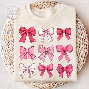 May include: A beige sweatshirt with a design of nine pink bows in different styles. The bows are arranged in three rows of three.