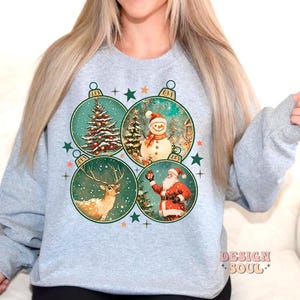 May include: A grey sweatshirt with a Christmas design featuring four baubles. Each bauble has a different image: a snowman, a deer, a Christmas tree, and Santa Claus. The baubles are surrounded by stars and snowflakes.