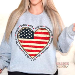 May include: Gray sweatshirt with a red, white, and blue heart-shaped American flag design. The words "Victory", "USA", "Freedom", "Independence", "Liberty", "Patriotism", "Unity", "Home of the Brave", "Land of the Free", and "America" surround the flag.