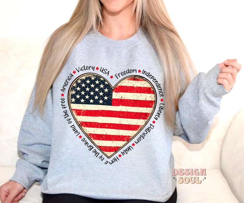 May include: Gray sweatshirt with a red, white, and blue heart-shaped American flag design. The words "Victory", "USA", "Freedom", "Independence", "Liberty", "Patriotism", "Unity", "Home of the Brave", "Land of the Free", and "America" surround the flag.